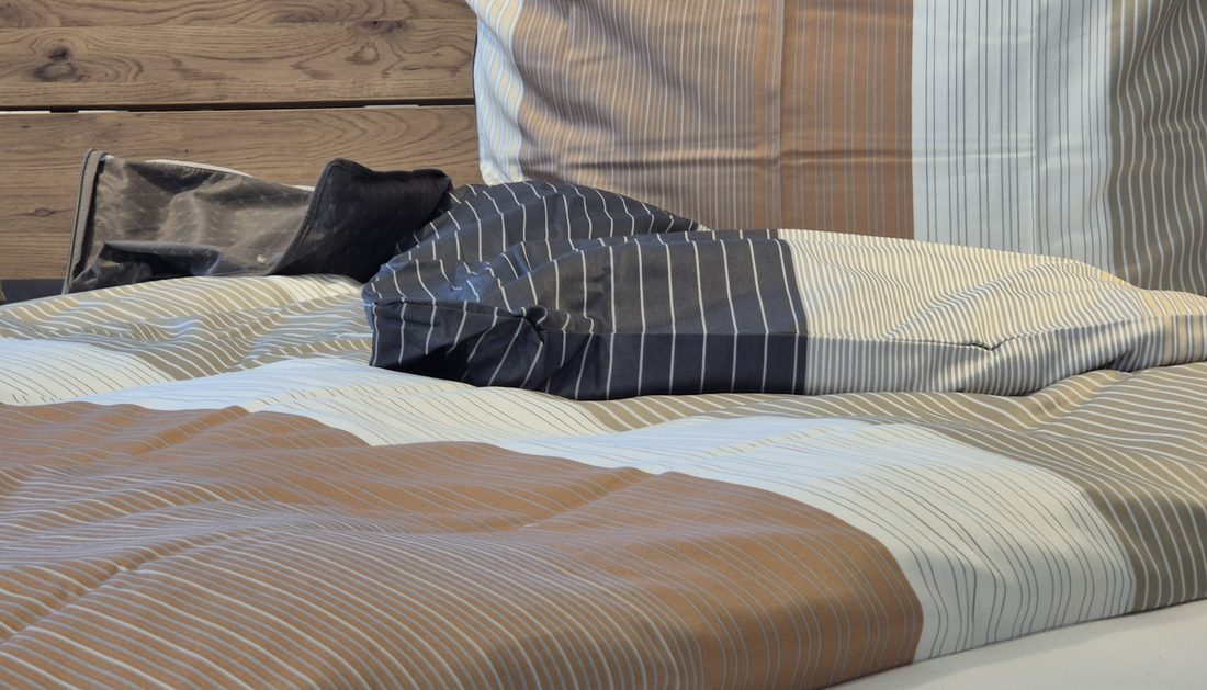 Bezogenes Bett im Showroom, gestreifte Bettwäsche in Beige und Dunkelblau vor einem Kopfteil aus Holz.